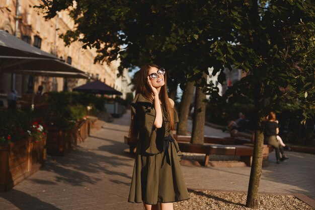 Portret van een vrolijke jonge vrouw in jurk aan te raken haar zonnebril op stedelijke achtergrond. Modieus meisje dat met mooi bruin haar aan camera glimlacht.