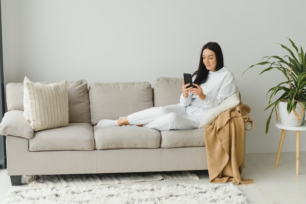 Portret van een vrolijke jonge vrouw met behulp van mobiele telefoon terwijl u ontspant op een bank thuis