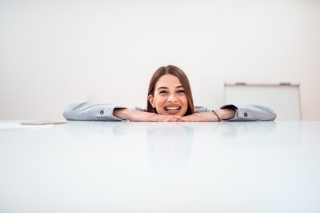 Portret van een vrolijke jonge vrouw met haar armen en hoofd op een witte tafel. Vooraanzicht, kopie ruimte.