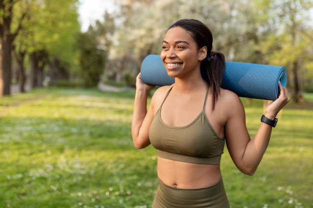 Portret van een vrolijke jonge zwarte vrouw die een sportmat op haar schouders houdt in de vrije ruimte van het stadspark
