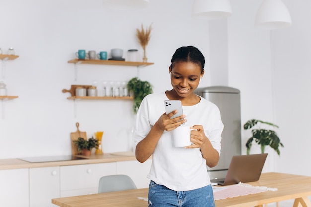 Portret van een vrolijke jonge zwarte vrouw die een telefoon vasthoudt en koffie drinkt in de keuken.