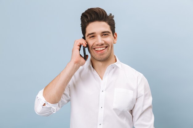 Portret van een vrolijke knappe jongeman permanent geïsoleerd op blauw, praten over de mobiele telefoon