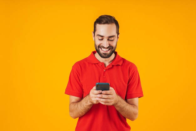 Portret van een vrolijke man in een rood t-shirt die lacht en een smartphone vasthoudt die over geel wordt geïsoleerd