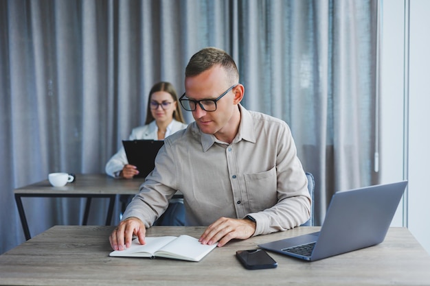 Portret van een vrolijke man IT-professional die werkt met een moderne laptop die aan een tafel zit en tijdens een pauze naar de camera glimlacht, een gelukkige menselijke programmeur in een bril voor oogcorrectie