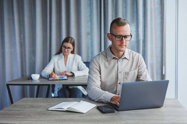 Portret van een vrolijke man IT-professional die werkt met een moderne laptop die aan een tafel zit en tijdens een pauze naar de camera glimlacht, een gelukkige menselijke programmeur in een bril voor oogcorrectie