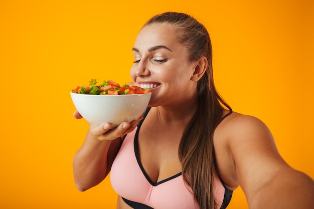 Portret van een vrolijke overgewicht fitness vrouw, gekleed in sportkleding staande geïsoleerd over gele muur, een selfie met mobiele telefoon nemen, kom met salade tonen