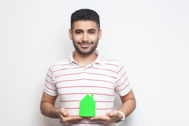 Portret van een vrolijke positieve, blije, optimistische jonge volwassen man in een t-shirt heeft een droom en houdt een klein huis van papier vast en plant een nieuw huis om te kopen. Binnen, geïsoleerd, studio-opname, kopieer ruimte, witte achtergrond