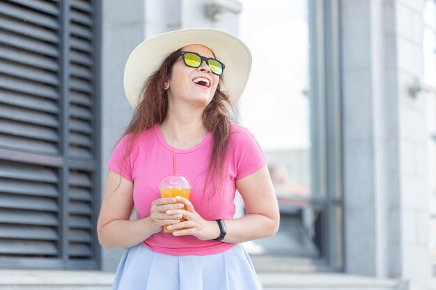 Portret van een vrolijke positieve jonge vrouw in een hoed en glazen met sap in haar handen lopen