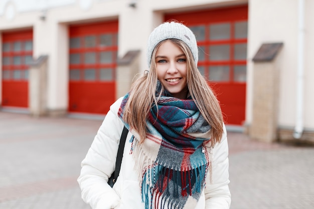 Portret van een vrolijke positieve jonge vrouw met een lieve glimlach met een warme stijlvolle geruite sjaal en een wit modieus jasje tegen de achtergrond van een gebouw met rode deuren. Het meisje glimlacht.