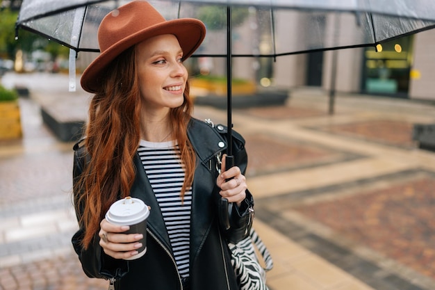 Portret van een vrolijke roodharige jonge vrouw met een modehoed die in de hand een kopje met hete koffie vasthoudt en zich met een transparante paraplu in de koude lenteregen bevindt