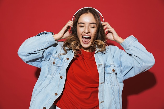 Portret van een vrolijke, stijlvolle jonge vrouw met een spijkerjasje dat geïsoleerd over de rode muur staat, naar muziek luistert met een koptelefoon, danst