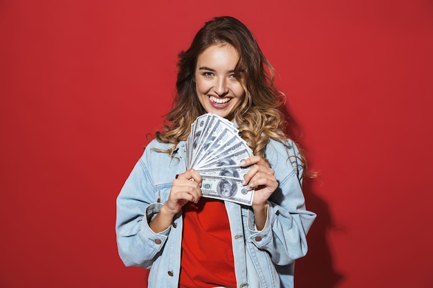 Portret van een vrolijke, stijlvolle jonge vrouw met een spijkerjasje dat geïsoleerd staat over een rode muur, poserend en geldbankbiljetten toont