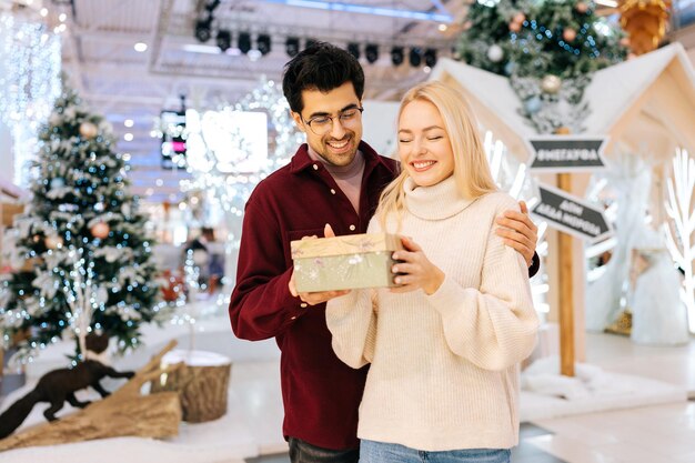 Portret van een vrolijke, vrolijke blonde vrouw die een geschenkdoos ontvangt van een liefhebbende echtgenoot die op kerstavond in de hal van het winkelcentrum Vieren staat