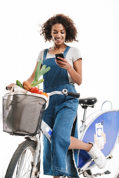 Portret van een vrolijke vrouw die een mobiele telefoon vasthoudt terwijl ze fietst met een boodschappentas die over een witte muur wordt geïsoleerd