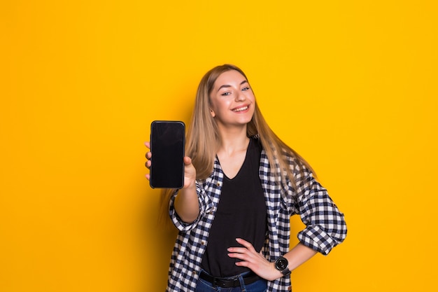 Portret van een vrolijke vrouw die het lege smartphonescherm over gele muur toont.