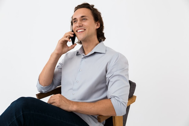 Portret van een vrolijke zakenman in een kantooroverhemd die op een smartphone praat terwijl hij in een fauteuil zit geïsoleerd over een witte muur