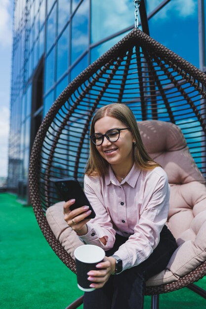 Portret van een vrolijke zakenvrouw in glazen die op een zonnige dag buiten een koffiepauze geniet van een succesvolle vrouwelijke manager die aan de telefoon praat terwijl hij tegen de achtergrond van het kantoor zit