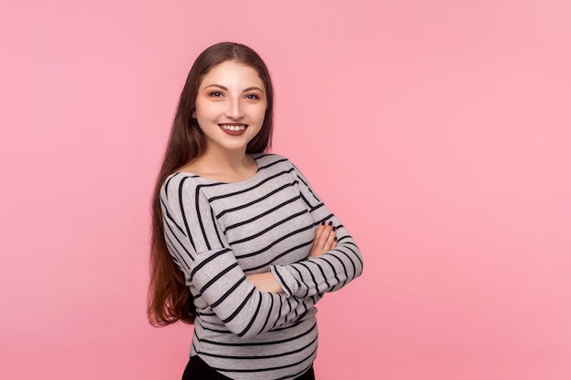Portret van een vrolijke zelfverzekerde vrouw in een gestreept sweatshirt die naar de camera kijkt met een brede glimlach, handen kruist en zich zelfverzekerd voelt, tevreden met het leven. studio-opname geïsoleerd op roze achtergrond