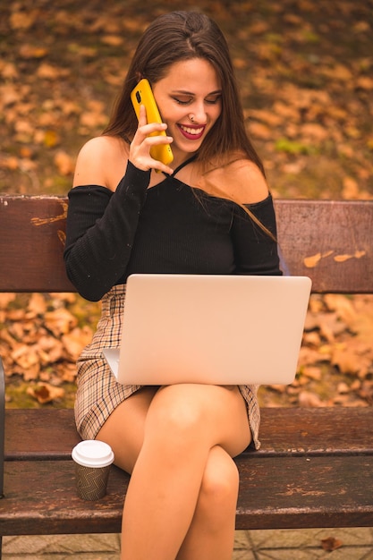 Portret van een vrouw in de herfst in een bos met bruine bladeren die naar de computer zit te kijken en telefoneert terwijl ze glimlacht