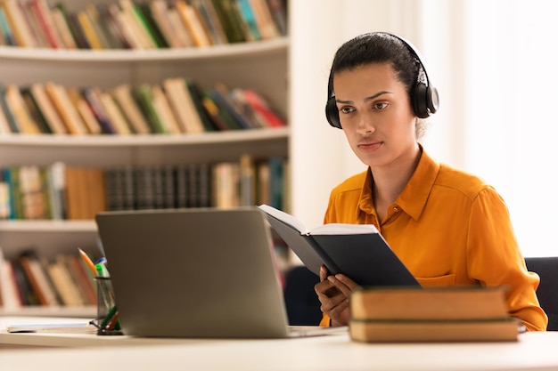 Portret van een vrouw in een draadloze hoofdtelefoon die een notitieboekje vasthoudt en naar een lezing kijkt op een laptop die aan tafel zit in de bibliotheek