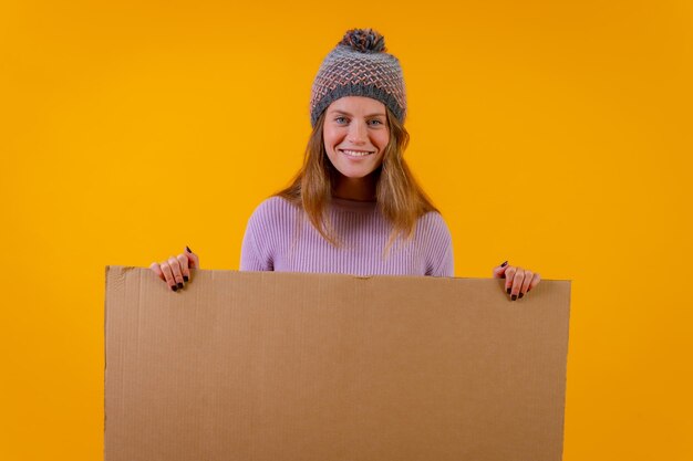 Portret van een vrouw in een wollen muts met een kartonnen bord op een gele achtergrond