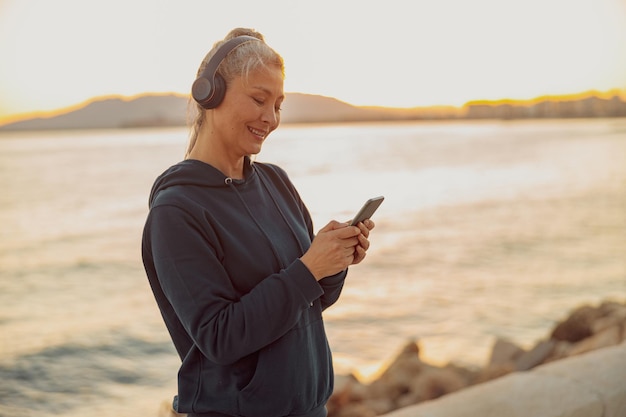 Portret van een vrouw in hoodie die bij de zee staat met smartphone