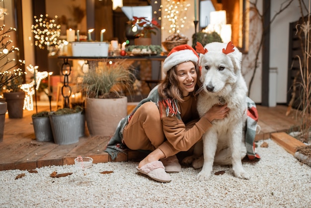 Portret van een vrouw in kerstmuts met haar schattige hond die een nieuwjaarsvakantie viert, samen zittend op het prachtig versierde terras thuis