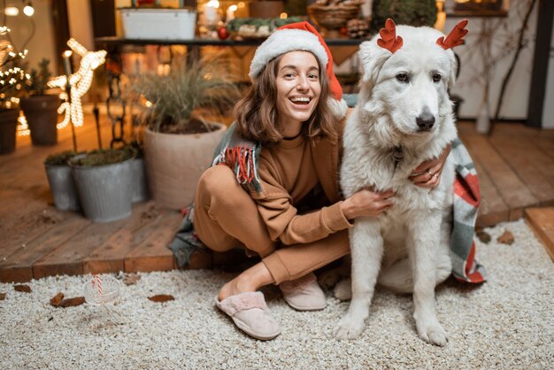 Portret van een vrouw in kerstmuts met haar schattige hond die een nieuwjaarsvakantie viert, samen zittend op het prachtig versierde terras thuis