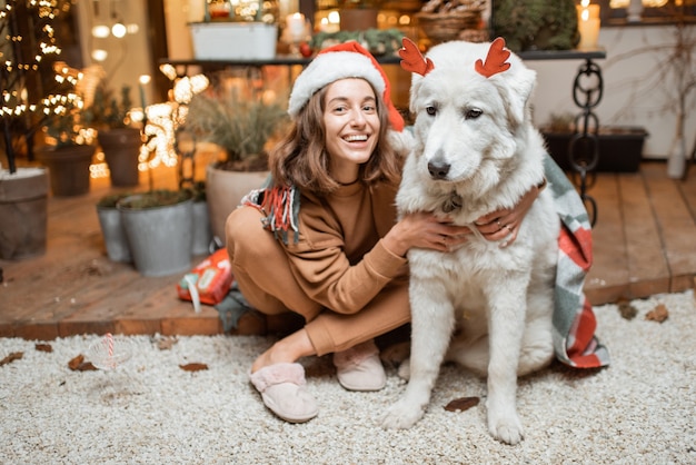 Portret van een vrouw in kerstmuts met haar schattige hond die een nieuwjaarsvakantie viert, samen zittend op het prachtig versierde terras thuis