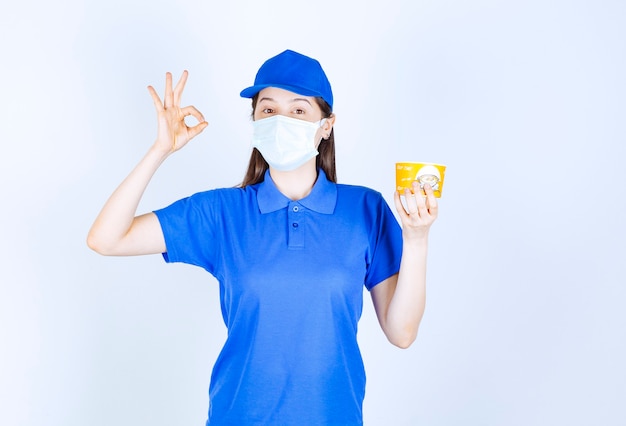 Portret van een vrouw in uniform en medisch masker met plastic beker die een goed gebaar toont