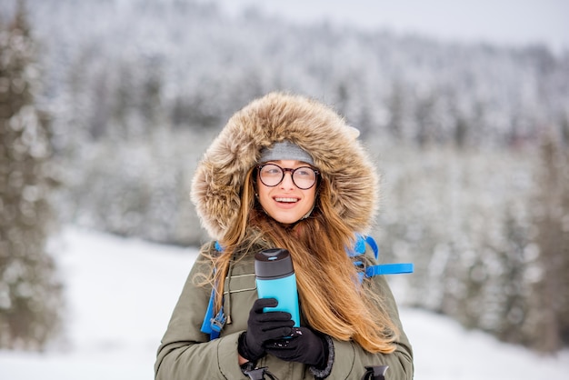 Portret van een vrouw in winterkleren die met rugzak, volgstokken en thermoskan wandelen in de besneeuwde bergen met bos op de achtergrond