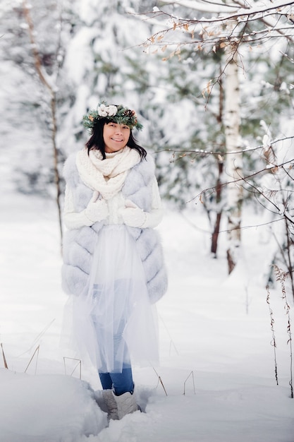 Portret van een vrouw in witte kleren in een koud winterbos. Meisje met een krans op haar hoofd in een besneeuwd winterbos