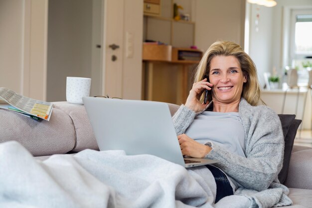 Foto portret van een vrouw met een mobiele telefoon terwijl ze met een digitale laptop op de bank zit