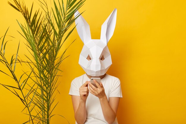 Portret van een vrouw met een wit T-shirt en een papieren konijnenmasker dat geïsoleerd over een gele achtergrond staat in de buurt van groen palmverlof met behulp van een mobiele telefoon die sms't met vrienden