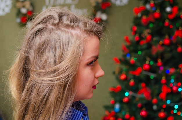 Portret van een vrouw met felrode lippen, blond lang haar tegen de nieuwjaarsboom. Jonge vrouw in een blauw herenoverhemd. Vakantie. Vrolijk kerstfeest. grappig gezicht