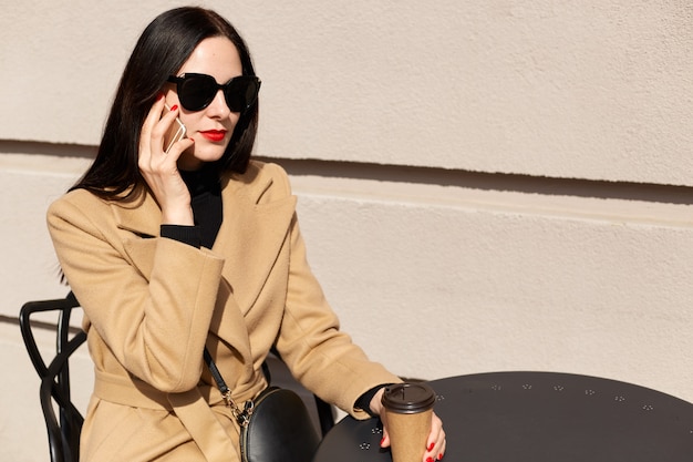 Portret van een vrouw met lang donker haar, het dragen van modieuze beige jas en afhaalmaaltijden koffie drinken