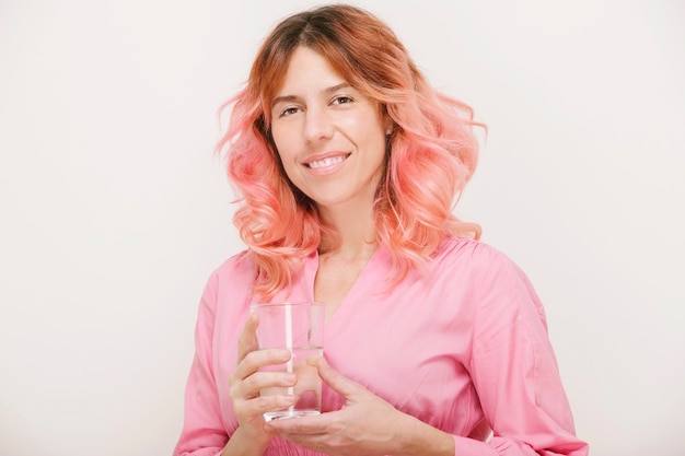 Portret van een vrouw met roze haar die met een glimlach naar de camera kijkt en een glas water vasthoudt