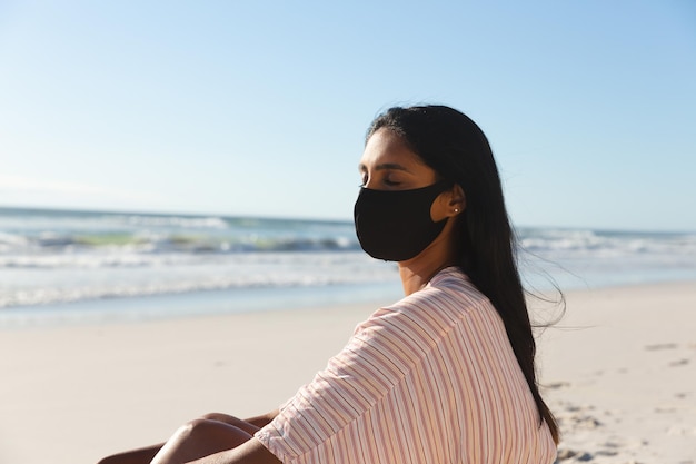 Portret van een vrouw van gemengd ras op strandvakantie met gezichtsmasker met gesloten ogen. vrije tijd buitenshuis vakantie aan zee tijdens coronavirus covid 19 pandemie.