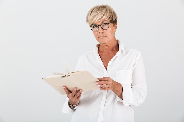 Portret van een vrouw van middelbare leeftijd met een bril die een boek vasthoudt en naar een camera kijkt die over een witte muur in de studio is geïsoleerd;