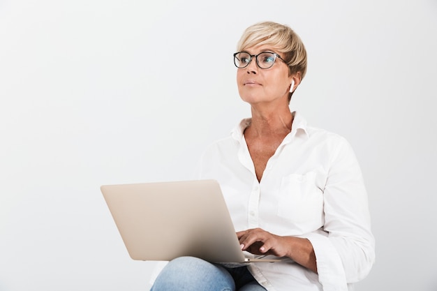 Portret van een vrouw van middelbare leeftijd met een bril en oordopjes die met een laptopcomputer zit geïsoleerd over een witte muur in de studio