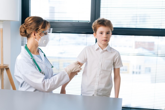 Portret van een vrouwelijke arts die een wit uniform draagt en pleister op de schouder van een kind aanbrengt na een vaccinatie-injectie bij het raam