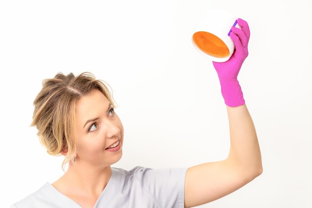 Portret van een vrouwelijke blanke schoonheidsspecialiste die een pot suikerpasta vasthoudt om te suikeren met roze handschoenen op een witte achtergrond