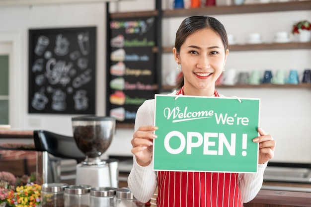 Portret van een vrouwelijke eigenaar die bij de poort van haar coffeeshop staat met een open uithangbord