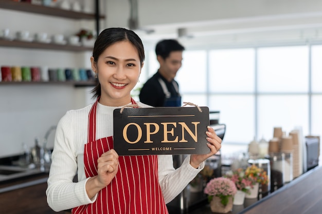 Portret van een vrouwelijke eigenaar die bij de poort van haar coffeeshop staat met een open uithangbord