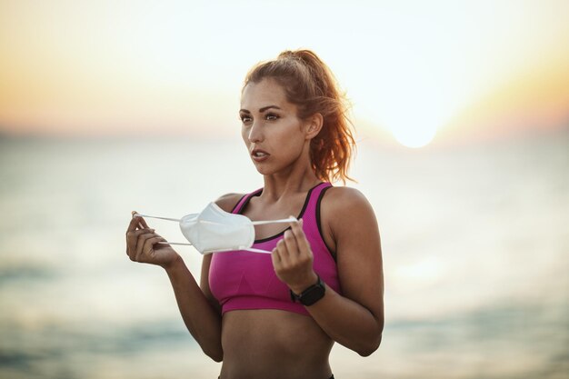 Portret van een vrouwelijke hardloper die een beschermend gezichtsmasker draagt tijdens een training in de buurt van het strand bij zonsondergang tijdens de COVID-19-pandemie.