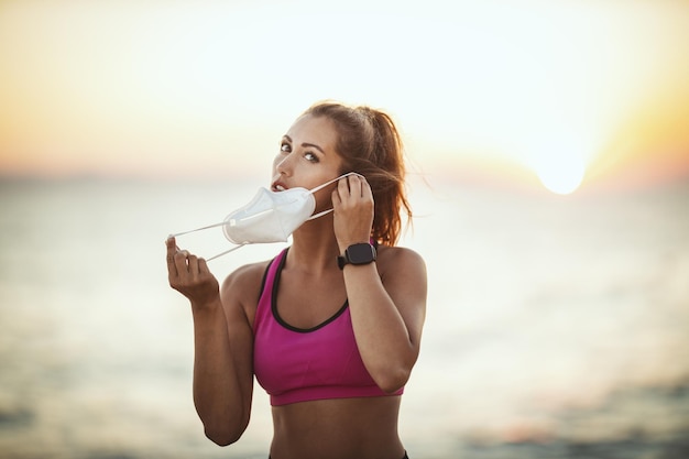 Portret van een vrouwelijke hardloper die een beschermend gezichtsmasker draagt tijdens een training in de buurt van het strand bij zonsondergang tijdens de COVID-19-pandemie.