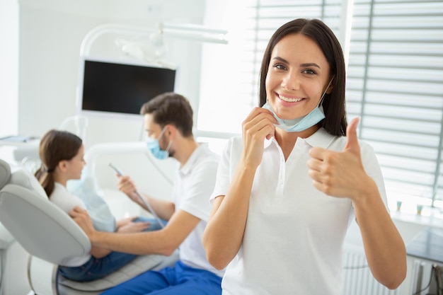 Portret van een vrouwelijke tandarts die duim toont terwijl haar collega met een kleine cliënt op de achtergrond werkt
