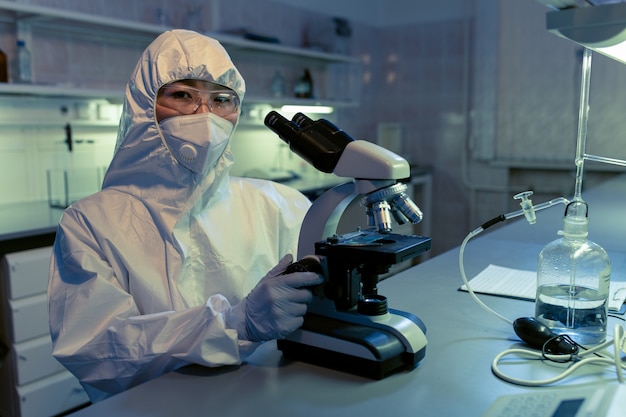 Portret van een vrouwelijke wetenschapper in beschermende werkkleding die naar de camera kijkt tijdens het werken met een microscoop in een chemisch laboratorium