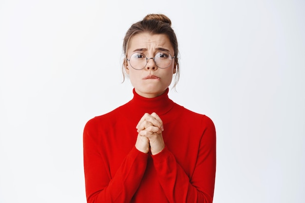 Portret van een wanhopig blond meisje dat om verontschuldiging smeekt of hulp nodig heeft, op de lip bijt en gebalde handen schudt in een smekend gebaar, om iets op wit te vragen