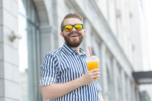 Portret van een wazig, stijlvol jong hipster-mannetje met baard en snor met vers sinaasappelsap in zijn handen. Gezond snackconcept.